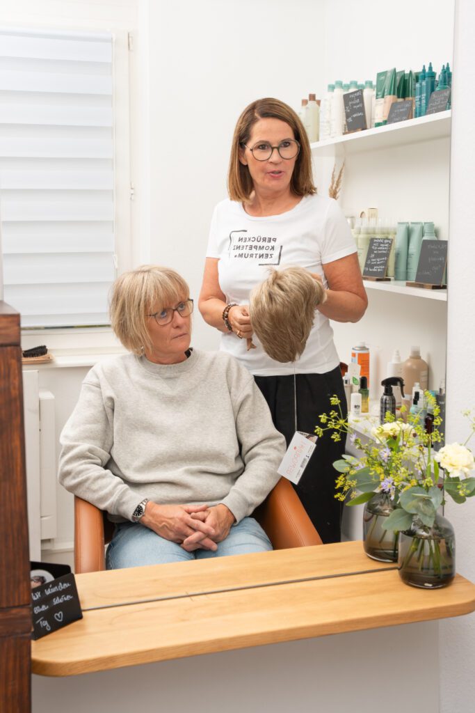 Beraterin Susanne Gress im weißen T-Shirt zeigt einer im Friseurstuhl sitzenden Kundin mit Brille eine blonde Perücke vor einem Spiegel