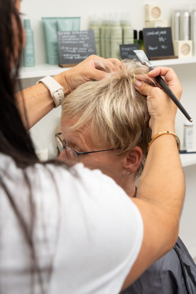 Eine Friseurin teilt das kurze blonde Haar der Perücke einer Kundin am Hinterkopf, um die Passform zu untersuchen. Sie hält ein Schneidewerkzeug in der Hand. Im Hintergrund stehen Regale mit Pflegeprodukten und Preisschildern